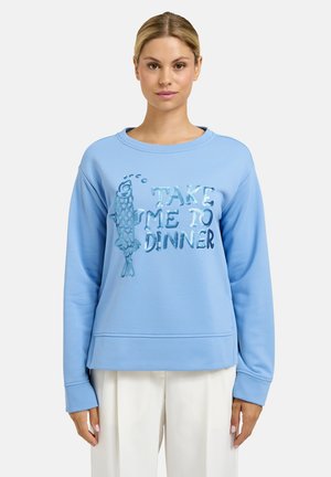 Woman wearing light blue sweatshirt with sequined fish and "Take Me to Dinner" text, paired with white pants, standing against plain background.