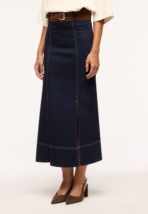 Donna con gonna midi di denim blu scuro con cuciture a contrasto marrone, cintura marrone, top bianco e scarpe con tacco marroni.