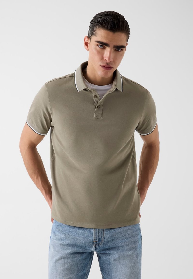 Jeune homme aux cheveux foncés coiffés en arrière, portant un polo beige avec un liseré blanc et un jean bleu clair, debout devant un fond uni.