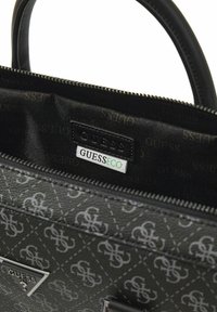 Schwarze, strukturierte Tasche mit Diamantmuster und silberner Hardware. Innenetikett mit der Aufschrift "GUESS" und "GUESS ECO". Reißverschluss sichtbar.