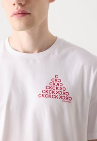 White cotton t-shirt featuring a red triangular logo design with repeated "CK" text and "Calvin Klein" branding at the bottom.