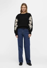 Pull noir avec des motifs floraux brodés sur les manches, associé à un jean droit bleu et des chaussures noires. Tissu lisse, coupe décontractée.