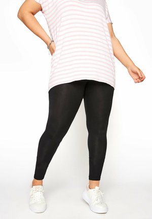 Czarne legginsy o gładkiej teksturze, w połączeniu z różowo-białą, prążkowaną, oversizową koszulą. Białe trampki dopełniają outfit.