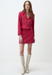 Veste courte en tweed rouge avec deux poches avant et boutons argentés, assortie à une mini-jupe, complétée par des bottes beiges montantes.