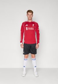 Maillot de football à manches longues rouge avec des accents blancs, arborant le logo de Liverpool FC et le sponsor. Shorts noirs et chaussettes blanches avec des rayures bleues.