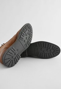 Zapatos de cuero marrón con una suela de goma negra texturizada que muestra un patrón de diseño detallado en la banda de rodadura. Vista en primer plano del diseño de la suela.