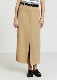 Personne portant une jupe midi beige avec une fente avant, une ceinture noire, un haut rayé, des mocassins noirs et des bagues aux doigts, se tenant devant un fond uni.