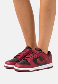 Baskets en cuir rouge et noir avec semelles blanches, avec des orteils perforés, un système de laçage à l'avant et le logo Nike sur le côté.