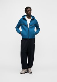 Zils rāvējslēdzēja džemperis ar melniem akcentiem, pieguļošs dizains un sānu kabatas, kombinēts ar vaļīgām melnām biksēm un gaiši pelēkiem sporta apaviem.