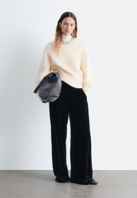 Flauschiger, hellpeachfarbener Pullover, darunter ein weißer Rollkragenpullover, dazu schwarze samtige weit geschnittene Hosen und eine dunkelgraue Handtasche. Schlichtes Design mit sanften Texturen.