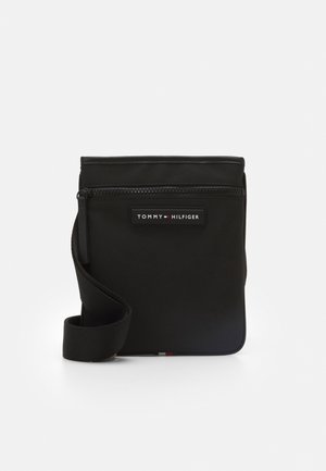 Sac bandoulière noir Tommy Hilfiger avec poche zippée à l'avant et sangle réglable sur fond blanc uni.