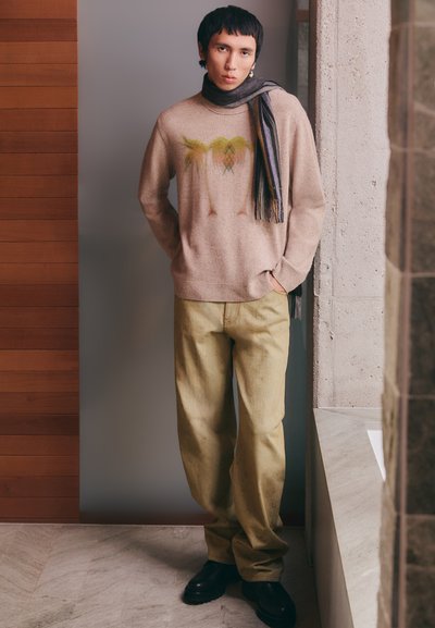Pull léger beige avec un motif de palmier, associé à un pantalon jaune pâle et une écharpe noire. Des chaussures noires élégantes complètent la tenue.