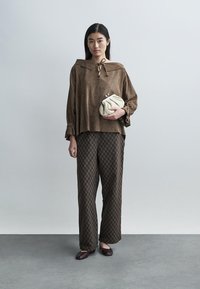 Top de camurça castanha com gola de laço, combinado com calças castanhas estampadas e uma carteira creme. Modelo em pé contra um fundo neutro.