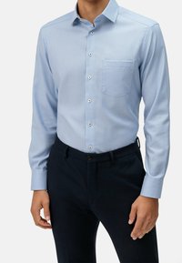 Světle modrá košile s dlouhým rukávem, límečkem na knoflíky, náprsní kapsou a jemnou texturou, kombinovaná s tmavými kalhotami.