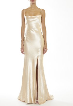 Femme portant une robe longue en satin couleur champagne avec fines bretelles et une fente haute sur la cuisse à l’avant, debout sur un fond blanc.