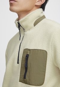 Fleece pullover in cream with a smooth olive green chest pocket; features a half-zip collar and black zipper detailing.