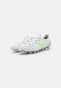 Scarpe da calcio bianche con accenti verdi, tomaia in pelle texturizzata, linee cucite e tacchetti sagomati per una maggiore trazione sulla suola.