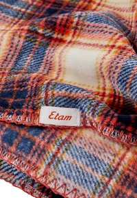 Koc w kratę w kolorach czerwonym, niebieskim i kremowym, wykonany z miękkiego materiału z obszytymi krawędziami i metką Etam w białym i pomarańczowym kolorze.