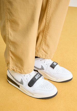 Weiße Nike-Sneaker mit schwarzen Riemen und gemustertem Swoosh, getragen zu beigen Hosen auf einem gelben Boden.