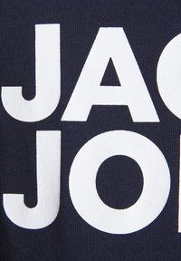 Mornarska tkanina z velikimi, bold belimi črkami, ki zapisujejo "JACK & JONES." Teksturirana površina z rebrastim vzorcem. Preprost, grafičen dizajn.