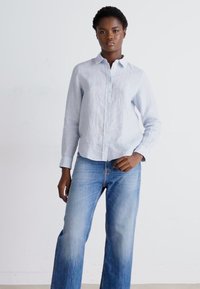 Jasnoniebieska lniana koszula z guzikiem i kołnierzykiem, noszona z wyblakłymi niebieskimi dżinsami w fasonie dzwonów. Gładka tekstura, standardowy krój oraz długie rękawy.