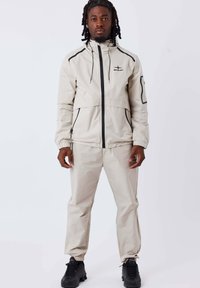 Giacca beige chiaro con accenti neri e zip, dotata di cappuccio e polsini elasticizzati, abbinata a pantaloni coordinati e sneakers nere.