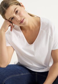 Hvid, kortærmet v-hals t-shirt i blødt stof, båret sammen med mørkeblå denim jeans. Modellen sidder og hviler sit hoved på den ene hånd.