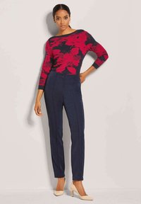 Rood gebloemd patroontrui met een bootnek, gecombineerd met donkerblauwe, op maat gemaakte broek. De outfit wordt compleet gemaakt met nude hoge hakken.