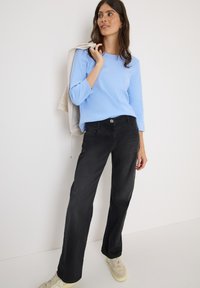 Lichtblauw langwerpig shirt, zwarte wijdvallende broek met voorzakken en crèmekleurige sneakers. Een grijze jas over de arm gedrapeerd.
