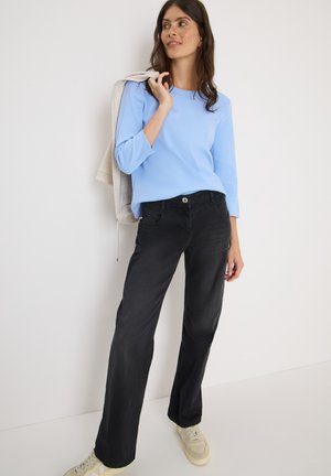 Haut à manches longues bleu clair, pantalon large noir avec poches avant et baskets crème. Une veste grise posée sur le bras.
