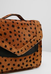 Ruskea käsilaukku, jossa mustia gepardinkuviota, läpällinen suljin ja pieni yläkahva, asetettuna yksinkertaisen harmaan ja valkoisen taustan eteen.