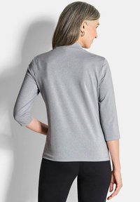 Top lungo a maniche con paillettes grigie, colletto alto e maniche a tre quarti, indossato con pantaloni neri, che mostrano una texture liscia e un design aderente.