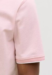 Nahaufnahme eines Arms einer Person, die ein hellrosa kurzärmliges Hemd aus strukturiertem Stoff mit einem doppellagigen Manschetten-Detail trägt.