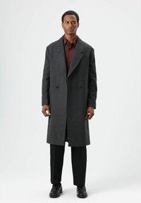 Antraciet wollen overjas met een dubbelgeknoopt ontwerp, brede revers en een gestroomlijnde silhouet, gecombineerd met donkere broek en zwarte schoenen.