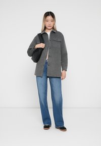 Grå ullskjorta med knappstängning och bröstficka, bärs över en vit topp. Blå vida jeans och svart väska kompletterar outfiten.