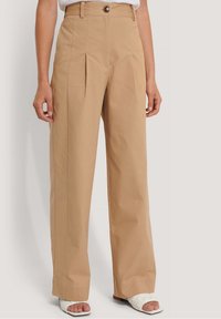 Plisowane, wysokie spodnie w jasnobrązowym materiale. Posiadają zapięcie na guzik, proste nogawki i gładką teksturę. W połączeniu z białymi sandałami na płaskiej podeszwie.