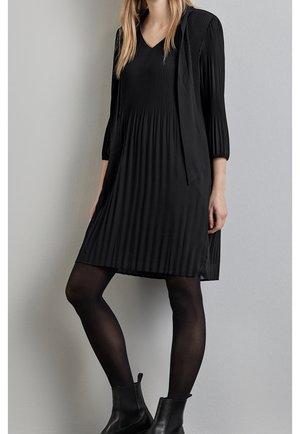 Czarna plisowana sukienka z dekoltem w kształcie litery V, rękawy do połowy przedramienia. Miękka tkanina, luźny krój, zestawiona z dopasowanymi czarnymi rajstopami i botkami za kostkę.