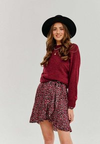 Burgundský sveter s dlhými rukávmi, skombinovaný s rozšírenou kvetinovou sukňou s ružovými a bielymi vzormi. Čierny klobúk s širokým okrajom dotvára vzhľad.