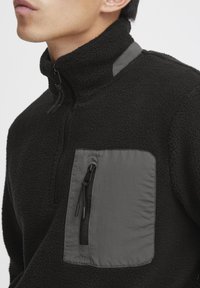 Schwarzer Fleece-Pullover mit hohem Kragen, ausgestattet mit einer grauen, mit Reißverschluss versehenen Brusttasche und einem glatten grauen Einsatz auf der Schulter. Reiche Textur.