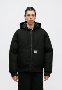 Črna kapucna jakna z zadrgo spredaj in velikimi žepi. Ima gladko površino, elastične manšete in kvadratno oznako na levi strani.