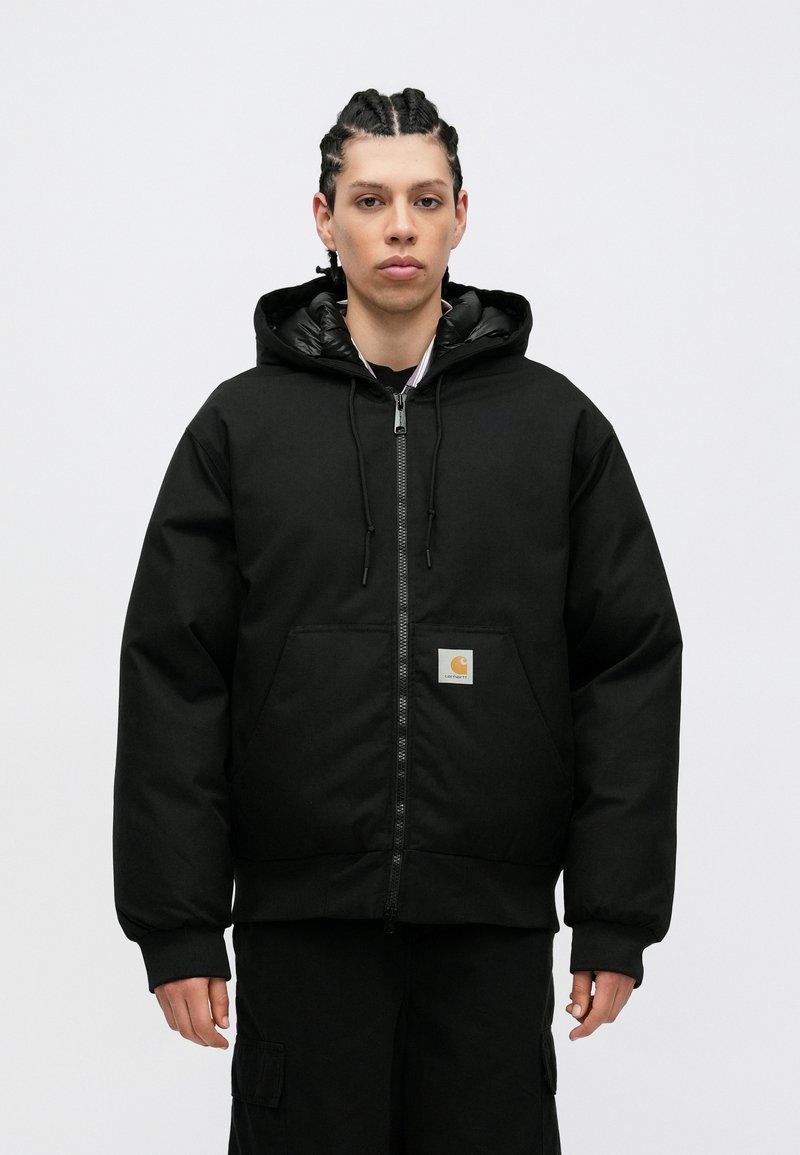 Chaqueta negra con capucha, cierre de cremallera en el frente y grandes bolsillos. Cuenta con un acabado suave, puños elásticos y un parche cuadrado con el logo en la parte izquierda.