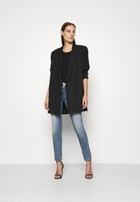 Svart dobbeltspent blazer med strukturerte skuldre, kombinert med en tettsittende svart topp og falmede blå skinny jeans. Åpen tå svarte hæler.