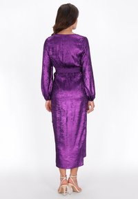 Mujer con un vestido midi de manga larga, brillante y color púrpura, con cintura fruncida, vista desde atrás, combinado con sandalias de tacón alto color nude.