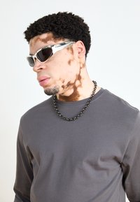 Sunglasses with a silver frame and dark lenses, worn with a gray long-sleeve shirt and a chunky black chain necklace.