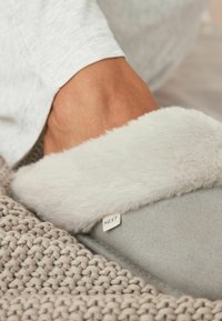 Gray slipper with a soft, white faux fur cuff, featuring a small "NEXT" tag. Textured, woven textile surface underneath.