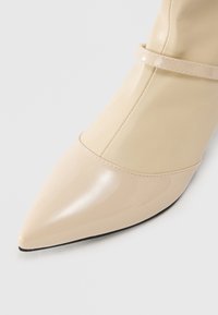 Beige pointed-toe boot with a patent finish on the toe and a matte finish on the upper, featuring a slim strap detail above the instep.