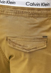 Bež pamuk kratke hlače s stražnjim džepom i pojasom s "Calvin Klein" logotipom u crnoj boji na bijeloj pozadini. Glatka tekstura, ležerni dizajn.