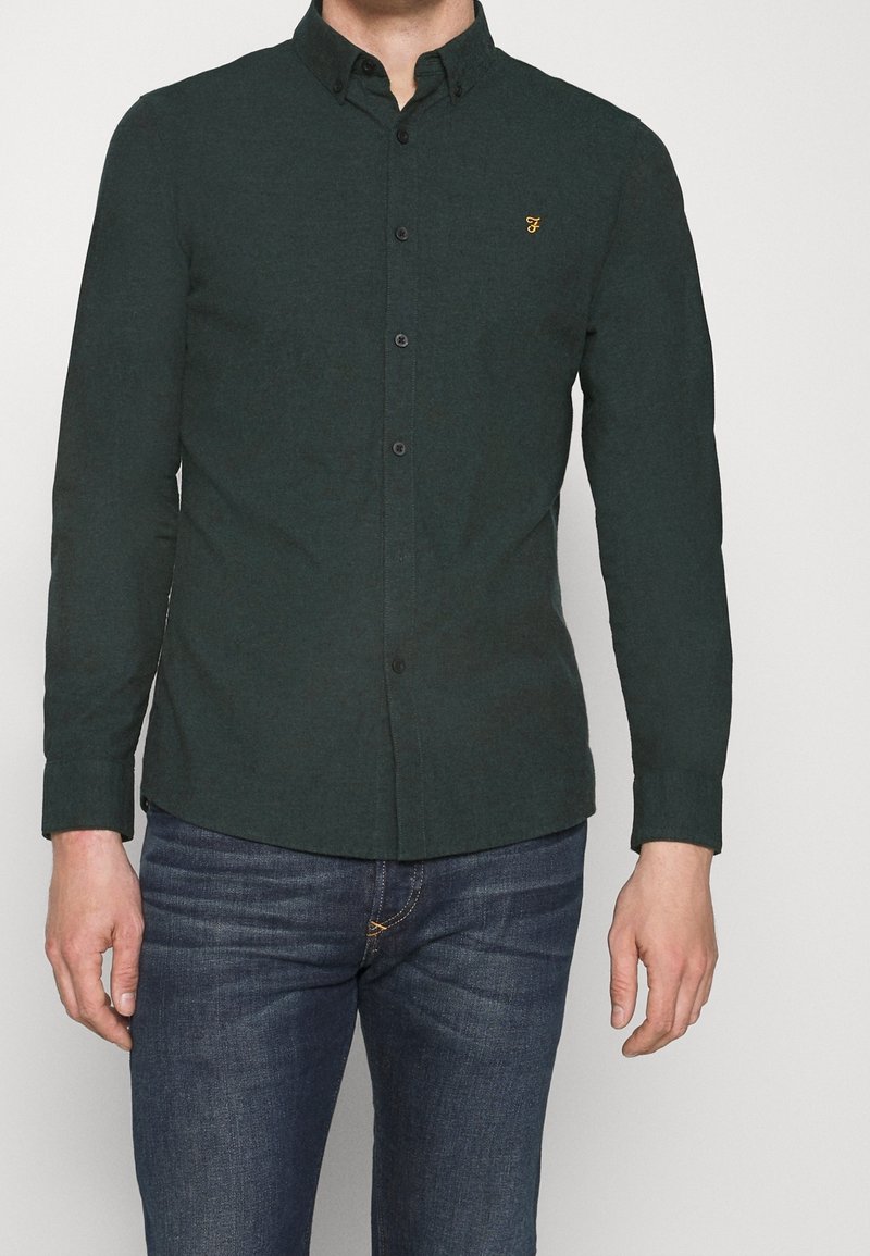 Camisa de manga larga de color verde oscuro con textura sutil, botones negros y un pequeño logo amarillo en el pecho izquierdo. Combinada con vaqueros azules.