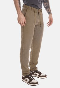 Pantaloni in cotone marrone chiaro con vita regolabile, tasche laterali e gambe leggermente affusolate. Indossati con sneakers nere e bianche.