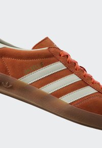 Narančasto-smeđi antilop tenisica s bijele tri pruge, gumeni potplat, narančaste vezice i natpis "GAZELLE" sa strane.